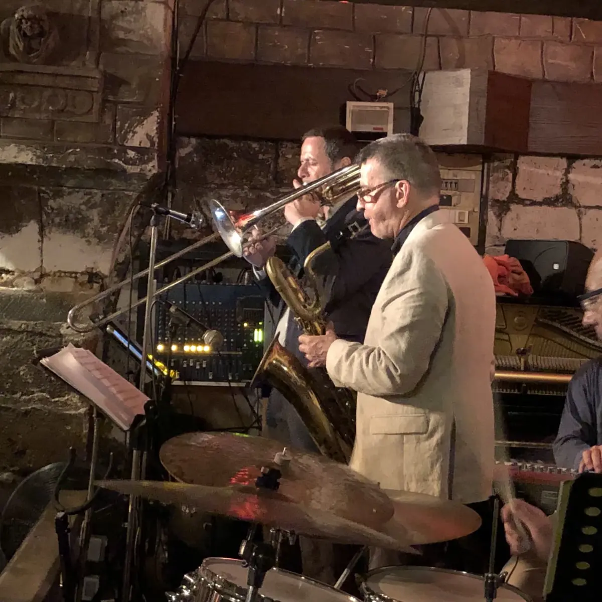 Two musicians play trombone and saxophone during an intimate jazz set in a dimly lit stone-walled venue.