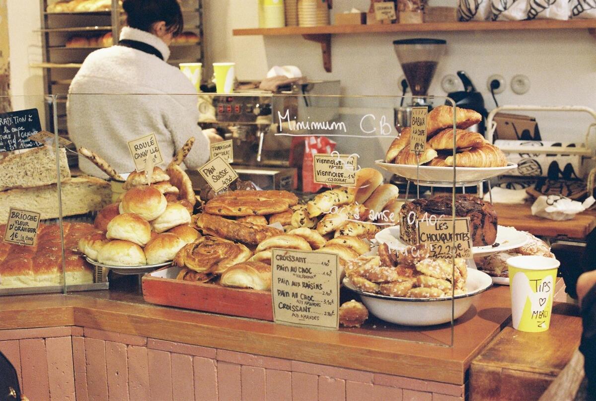 A well-stocked glass display case in a bakery, showcasing various golden French pastries like croissants, pain au chocolat, chouquettes, and large brioche, with handwritten price signs in French and "Minimum CB" text. A person is working in the background.