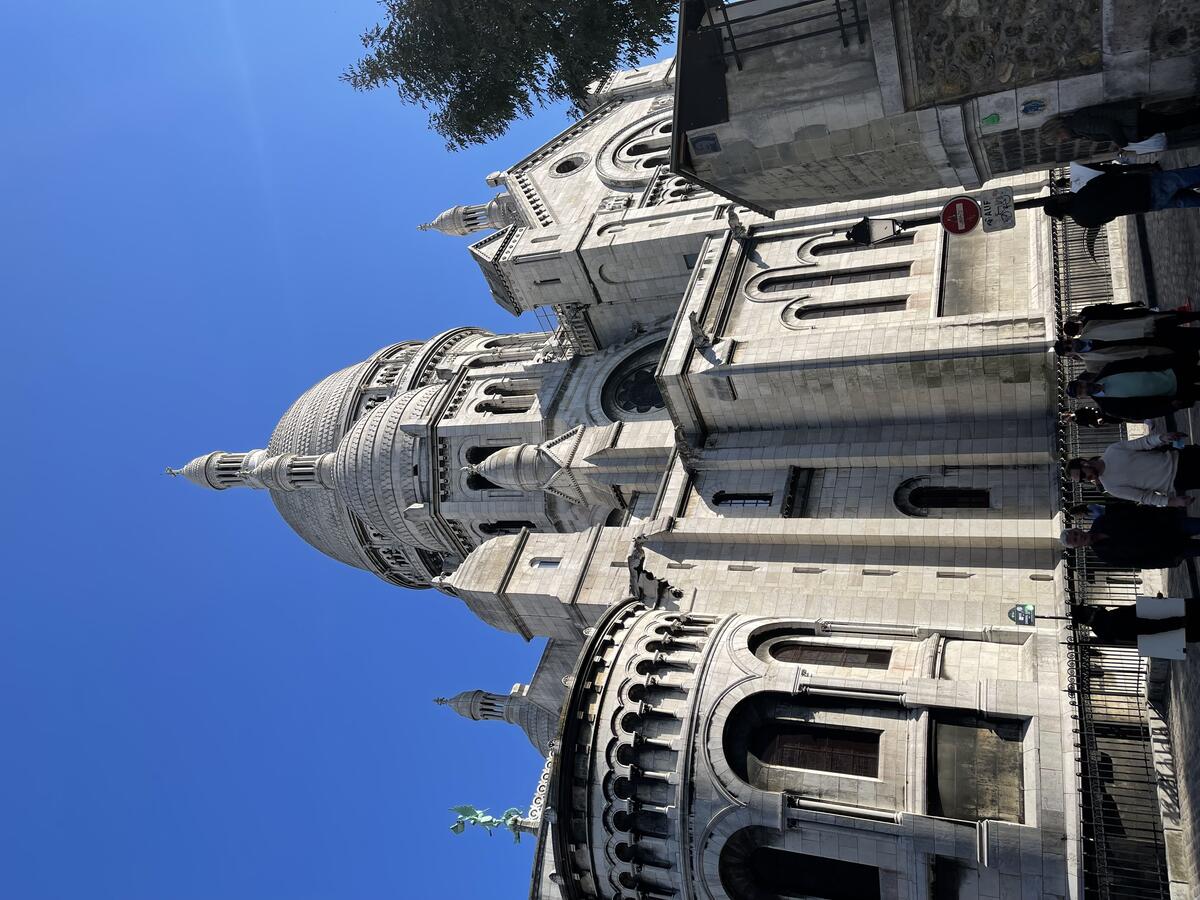 The large, white stone Sacré-Cœur Basilica in Paris stands under a clear blue sky, with its central dome and arcaded apse prominent, and people walking below.