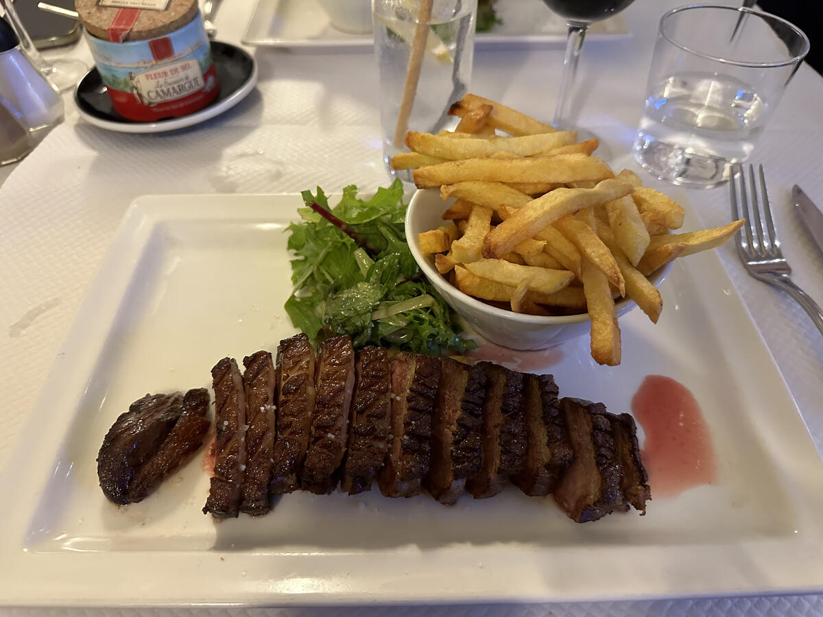 A white rectangular plate holds sliced grilled meat, a small bowl of golden french fries, and a side salad, set on a table with salt and water glasses.