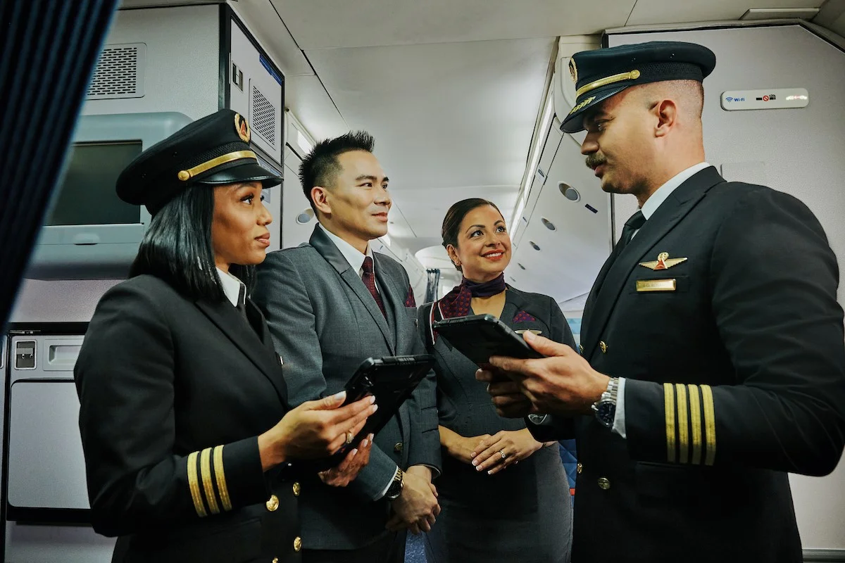 Two Delta pilots and two Delta flight attendants stand together in an airplane aisle, holding tablets and talking.