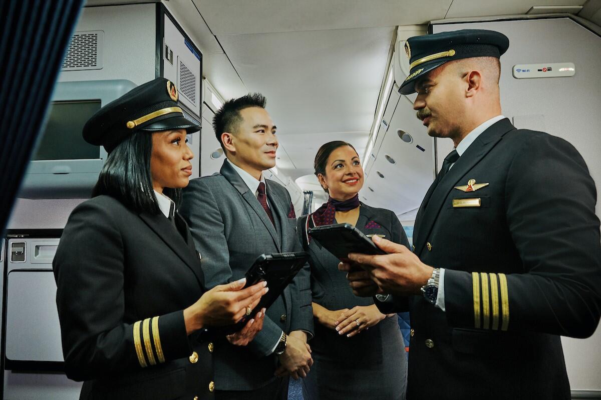 Two Delta pilots and two Delta flight attendants stand together in an airplane aisle, holding tablets and talking.