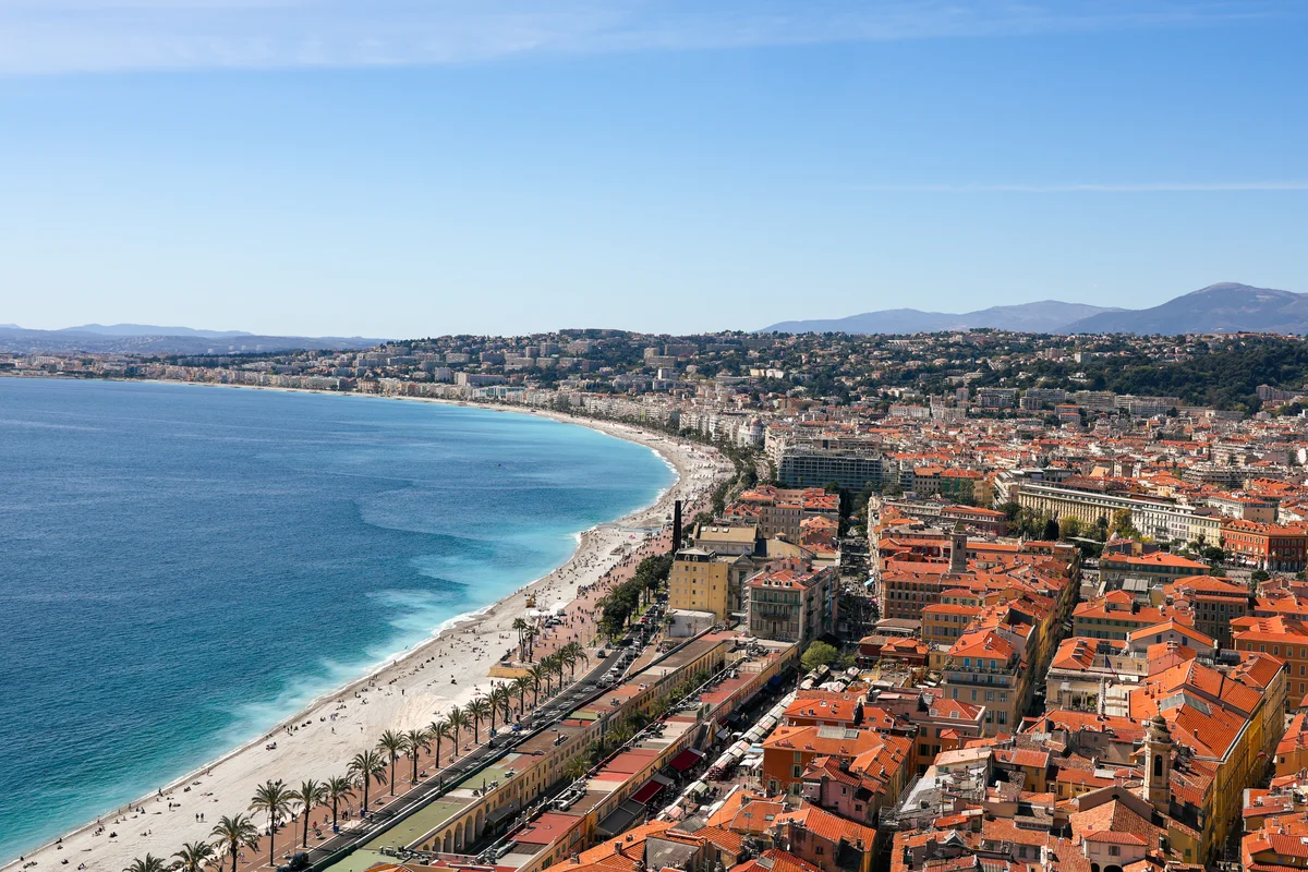 Aerial view of a curved Mediterranean beach and waterfront city with red-tiled roofs.