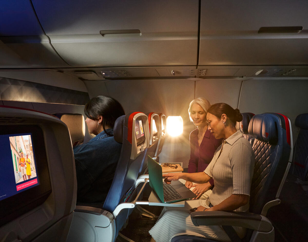 Two female Delta customers on board an aircraft working on laptops, with warm sunlight streaming through the windows.