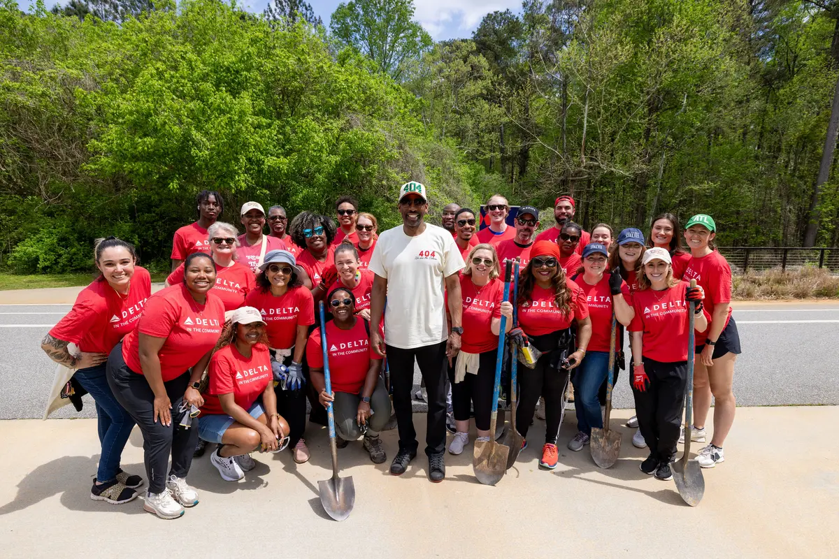 A large group of Delta volunteers wearing red “Delta in the Community” shirts stand together outdoors holding shovels, gathered on a paved area with trees in the background.