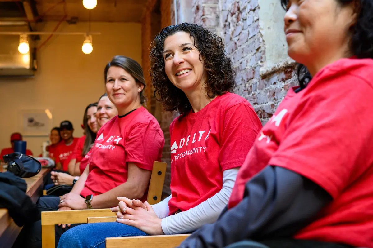 Delta volunteers wearing red “Delta in the Community” shirts sit side by side on chairs inside an indoor space with exposed brick walls during a community event.