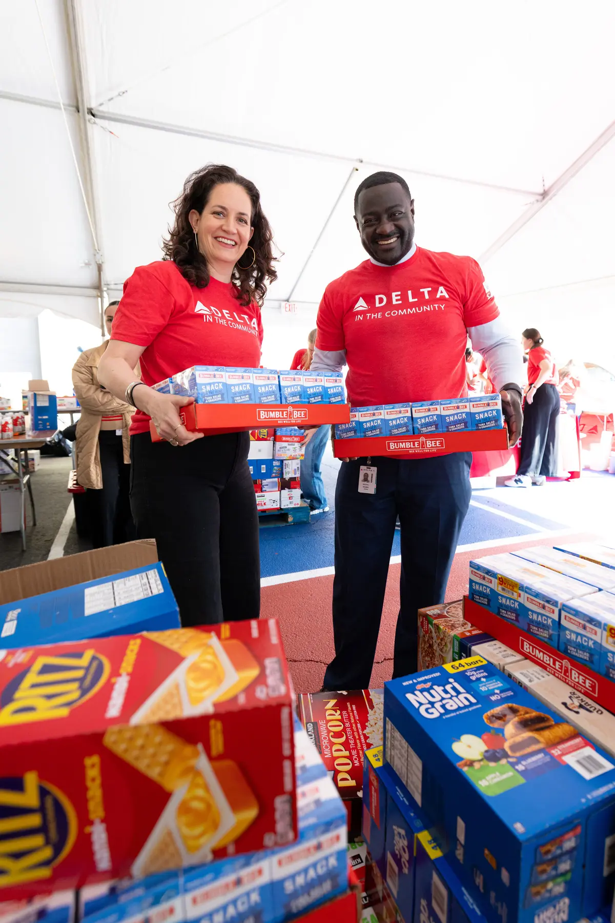 Two Delta volunteers wearing red “Delta in the Community” shirts carry boxes of snack food at a community packing event inside a large tent.