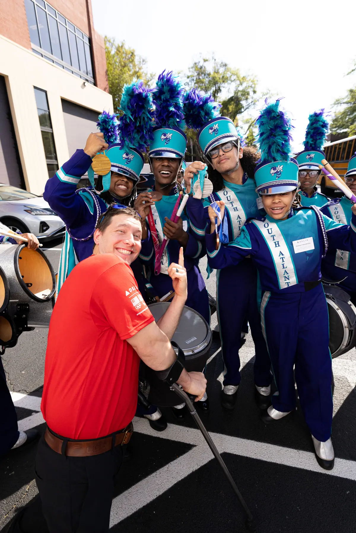 Delta volunteer in a red shirt interacts with a marching band in teal and purple uniforms, with drums and feathered hats, during an outdoor community event.