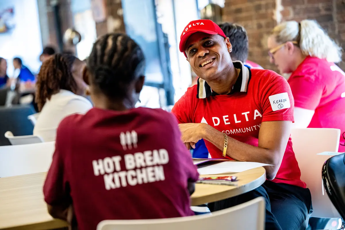 Delta volunteer in a red “Delta in the Community” shirt sits at a table speaking with a community member wearing a “Hot Bread Kitchen” shirt inside a bright indoor space.