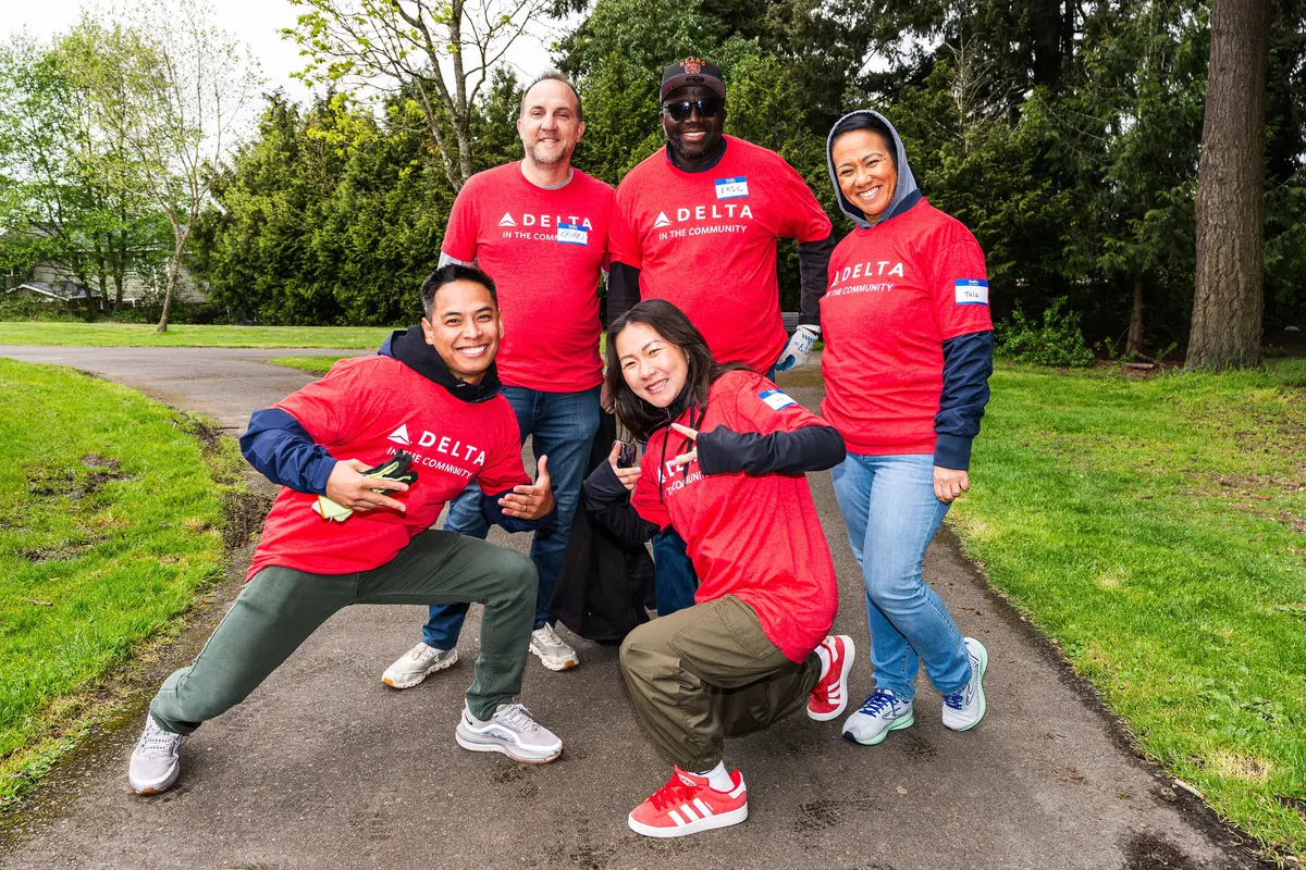 A group of Delta volunteers wearing red “Delta in the Community” shirts pose together on a park path, surrounded by green grass and trees.