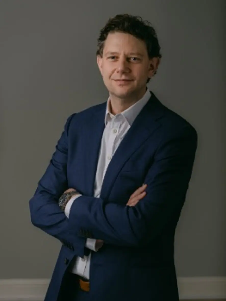 Studio portrait of a person in a dark suit and white shirt, standing with arms crossed against a plain gray background.
