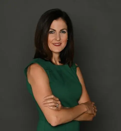 Studio portrait of a person with shoulder-length dark hair, wearing a sleeveless green dress and standing with arms crossed against a plain gray background.