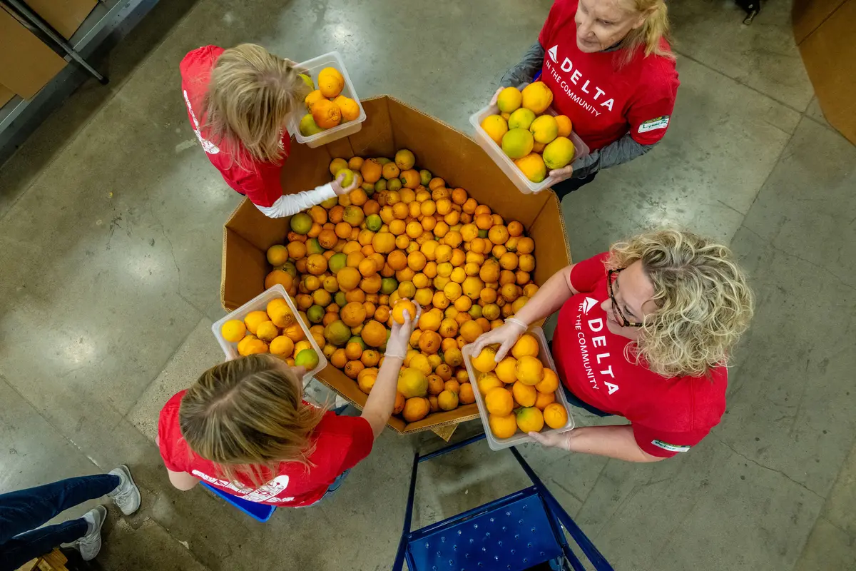 Overhead view of Delta volunteers wearing red “Delta in the Community” shirts sorting oranges into containers around a large bin inside a warehouse setting.