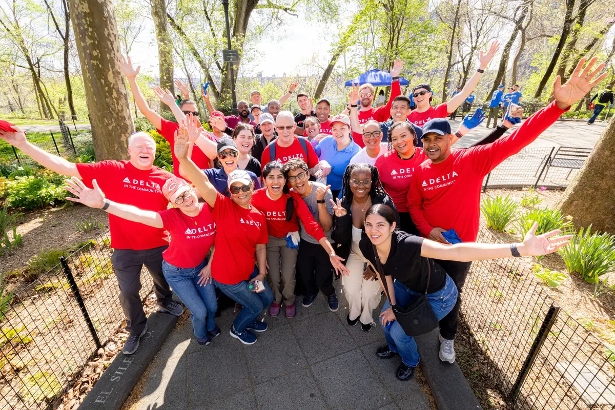 A large group of Delta volunteers wearing red “Delta in the Community” shirts stand together in a park with arms raised, gathered along a paved path surrounded by trees.