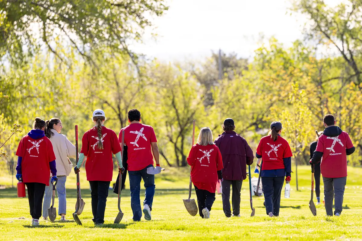 A group of Delta volunteers wearing red shirts walk across a grassy park carrying shovels, heading to a tree‑planting activity on a sunny day.