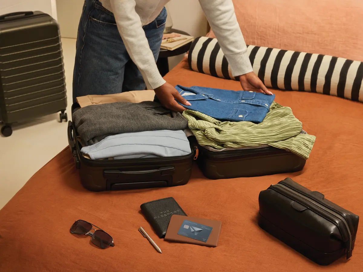 Person packing clothes into an open suitcase on a bed, with a Delta SkyMiles American Express Card, wallet, passport, and sunglasses placed nearby.