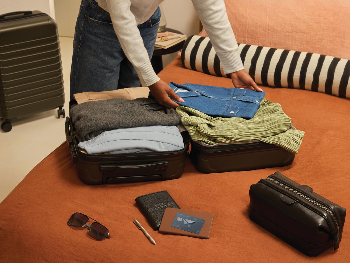 Person packing clothes into an open suitcase on a bed, with a Delta SkyMiles American Express Card, wallet, passport, and sunglasses placed nearby.