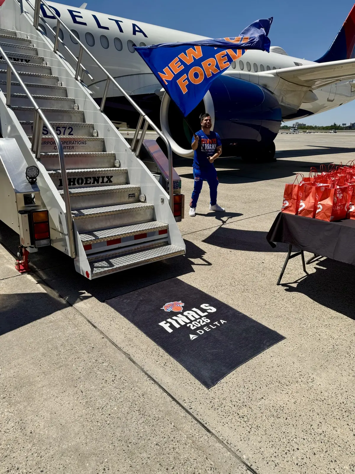 Orange New York Knicks foam finger reading “#1 FAN” sits atop Delta-branded blue towels at an airport display, with travelers and check-in counters in the background.