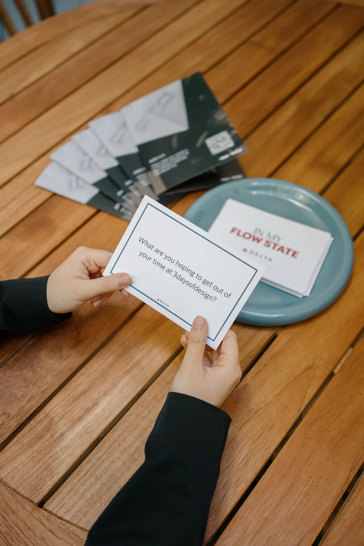 Hands holding a prompt card over a wooden table beside a bowl of cards, reading “What are you hoping to get out of your time at 3daysofdesign?”