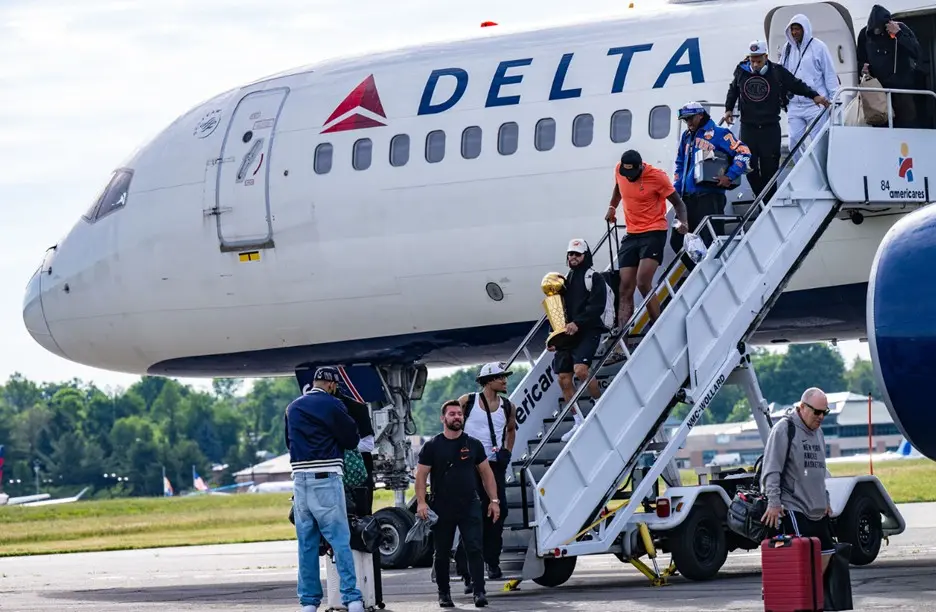 People exit a Delta airplane via stairs on the tarmac, with one person carrying a gold trophy while others hold bags nearby.