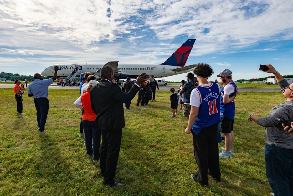 Spectators stand on grass watching a Delta airplane taxi on the runway at an airport.