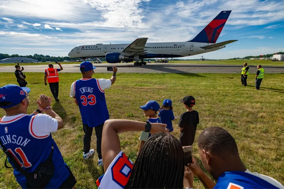 Group of people in sports jerseys standing on grass watching and taking photos of a Delta airplane taxiing on a runway while staff in safety vests stand nearby.
