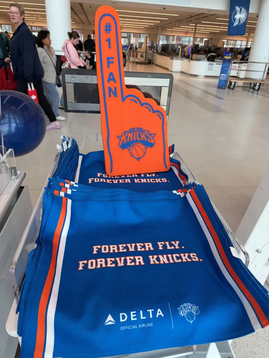 Bright orange New York Knicks foam finger displayed on a stand of blue Delta‑branded rally towels reading “Forever Fly. Forever Knicks” in an airport terminal, with travelers and security area in the background.