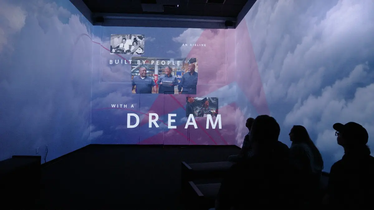 Silhouetted viewers watch an immersive projection of clouds with photos of people and the text “BUILT BY PEOPLE WITH A DREAM.”