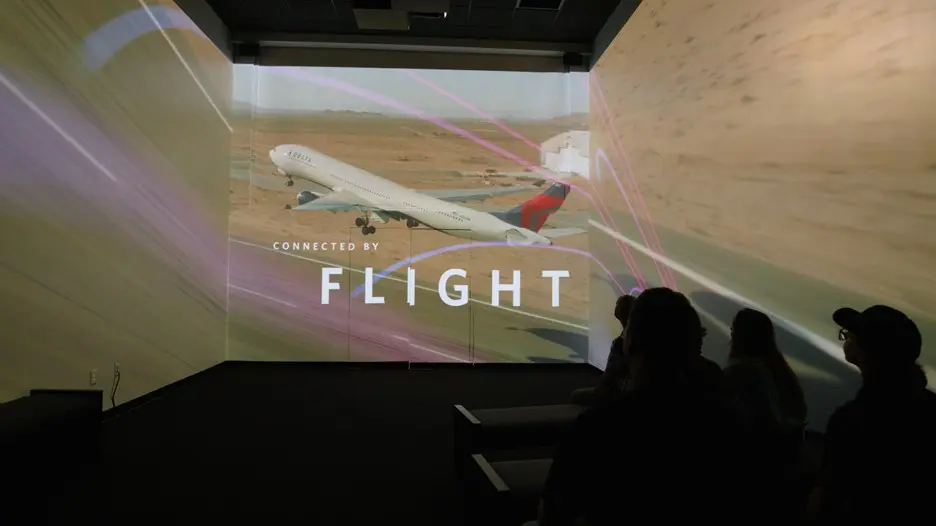 Silhouetted audience in a dark room viewing a multi-wall projection of a plane taking off, with the text “CONNECTED BY FLIGHT” overlaid.