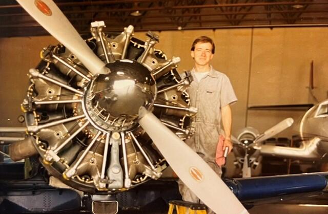 Person in a maintenance jumpsuit standing beside a vintage radial aircraft engine with a large propeller in a hangar.