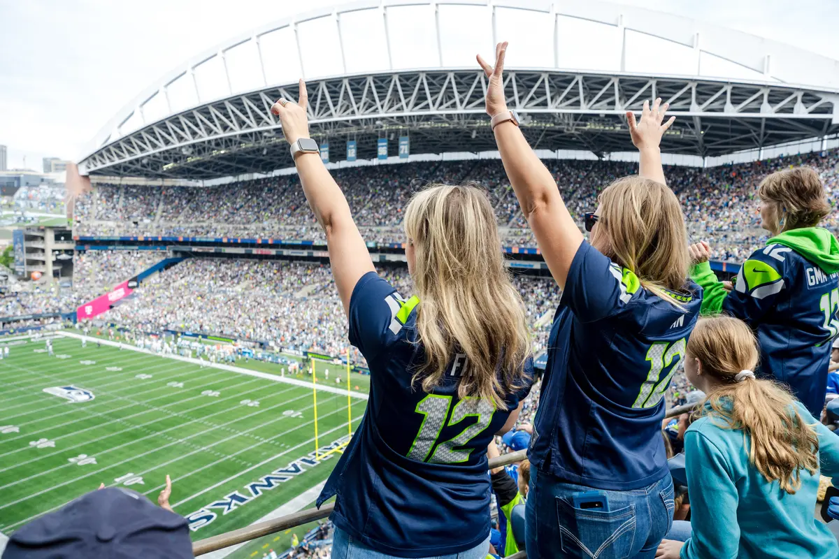 Fans wearing Seattle football jerseys raise their arms while watching a game from the stands of a crowded stadium.