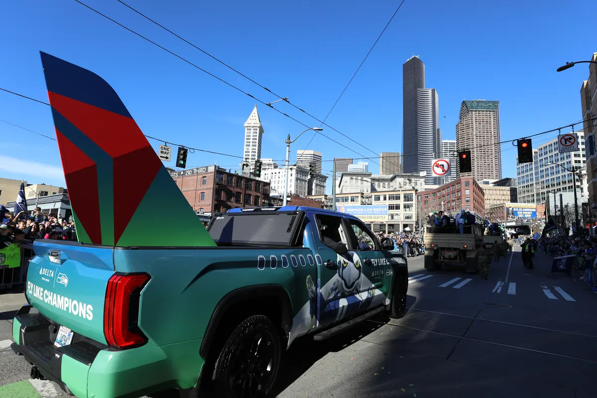 Delta-branded vehicle carrying a large airplane tail design during a parade through downtown Seattle, with crowds lining the streets and the city skyline in the background.