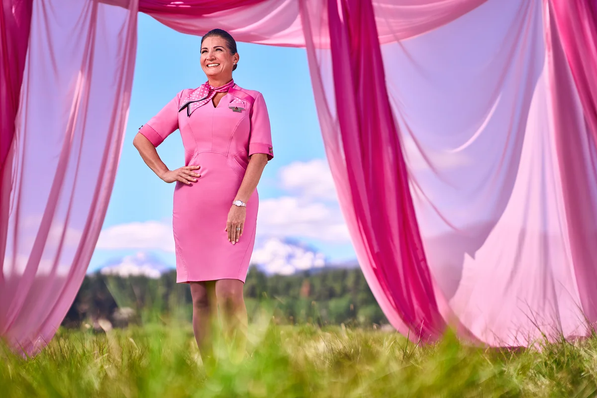 Person in a pink uniform dress standing outdoors between flowing pink fabric panels, with grass, trees and mountains in the background.