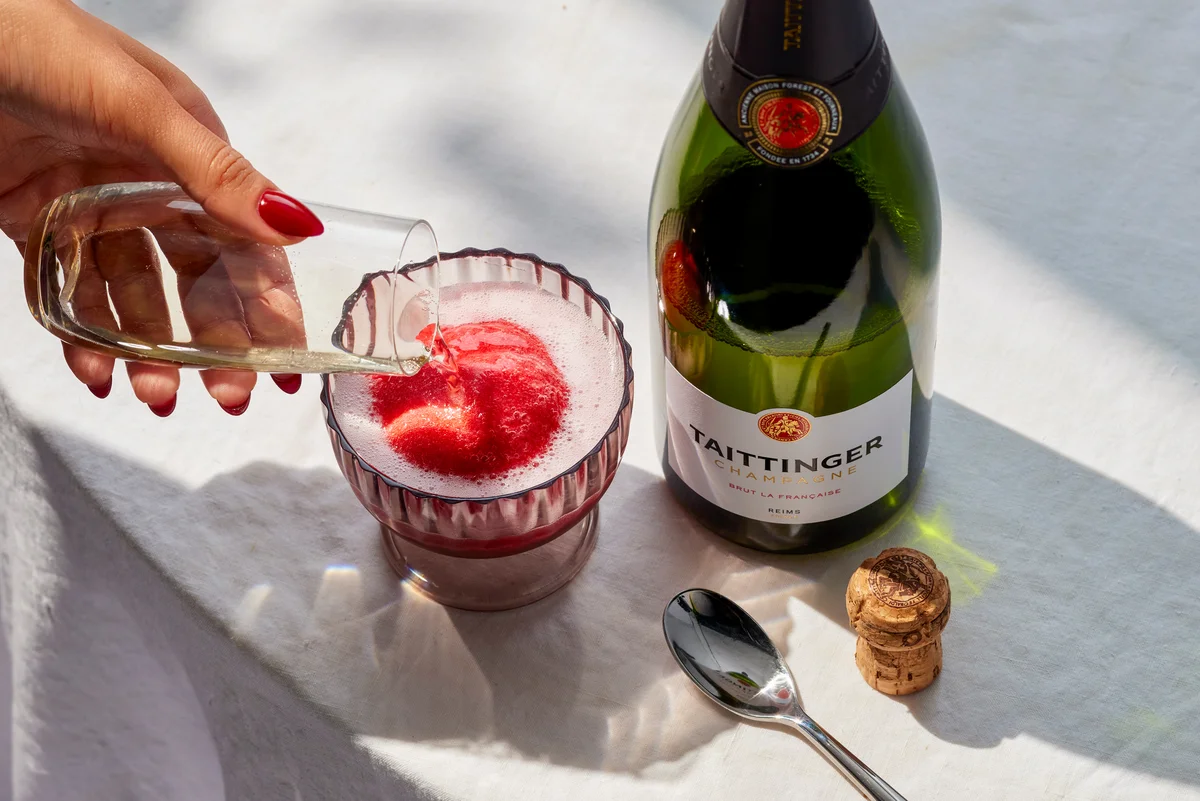 A hand pours sparkling wine over a red fruit sorbet dessert beside a champagne bottle on a white tablecloth.