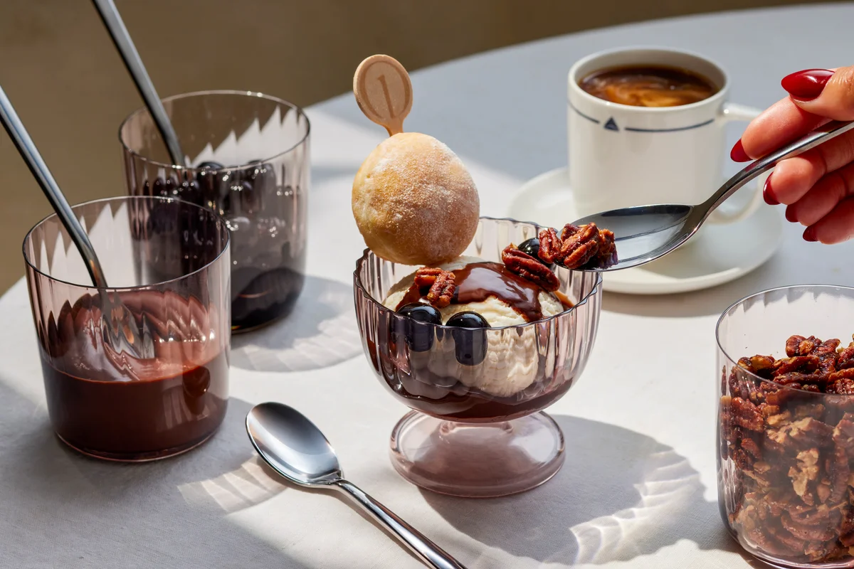 Ice cream sundae topped with chocolate sauce, pecans, and cherries, served with coffee and dessert accompaniments on a table.