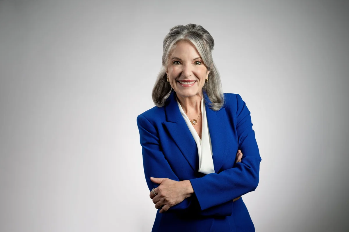 Studio portrait of a person wearing a blue blazer and white blouse, standing with arms crossed against a light gray background.