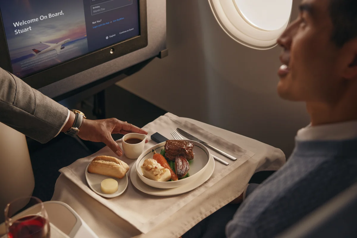 Flight attendant serves a plated meal with bread and vegetables to a passenger in a Delta One cabin.