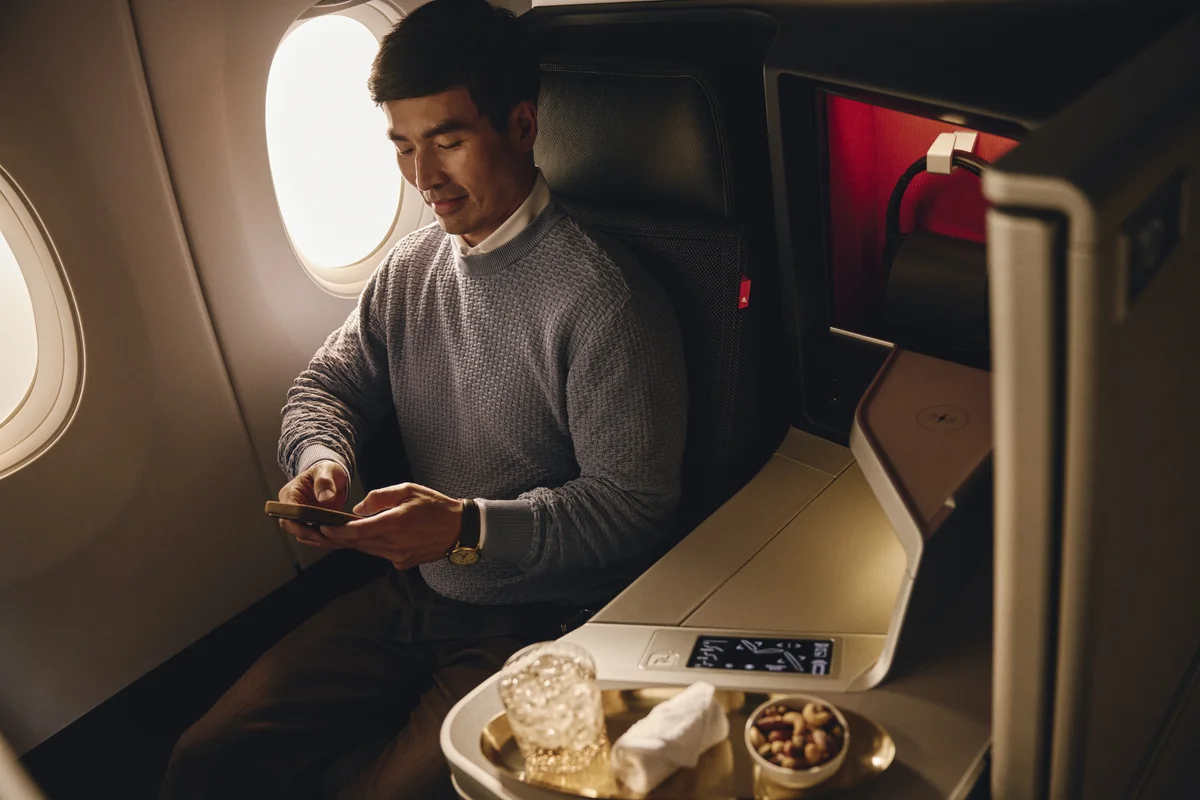 Customer seated in a premium airline suite, using a mobile phone beside a tray with a beverage and snacks.