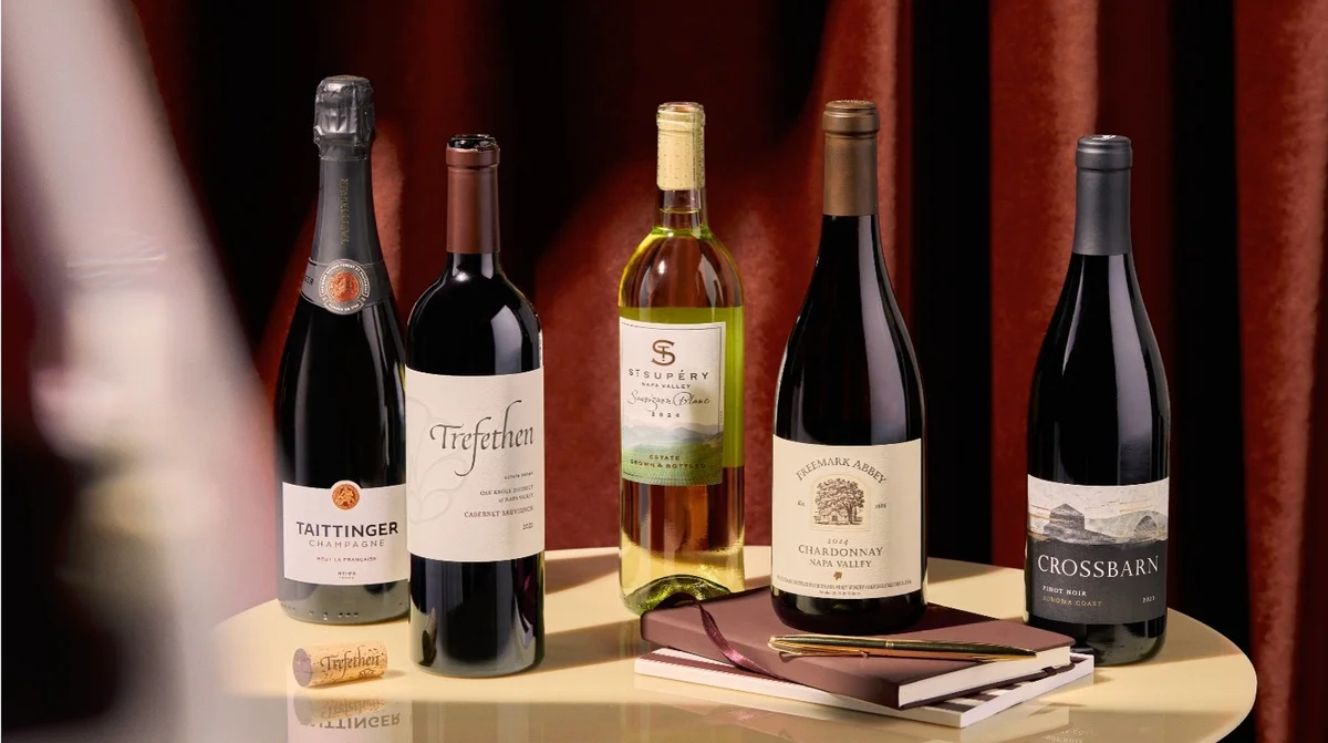 Five wine bottles arranged on a table with notebooks and a pen, set against a rich red curtain backdrop.