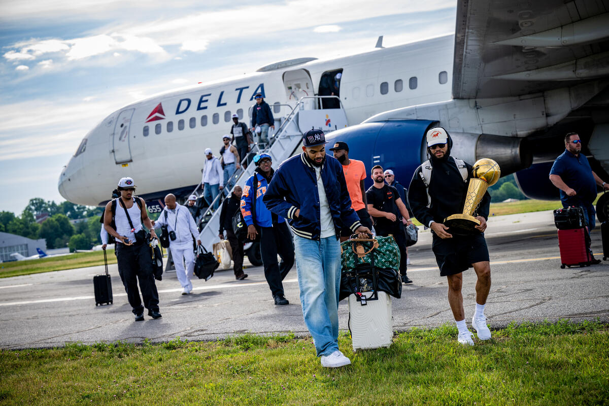 Passengers walk across a grassy airport runway after disembarking a Delta airplane, with one person carrying a large gold trophy.
