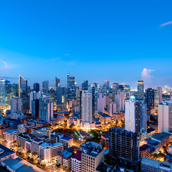 A panoramic view of a modern city skyline at dusk, with illuminated high-rise buildings, busy streets, and a bright blue evening sky.